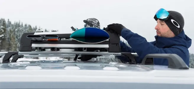 Man in a beanie and ski goggles loading skis into a roof-mounted ski rack, snowy trees behind him