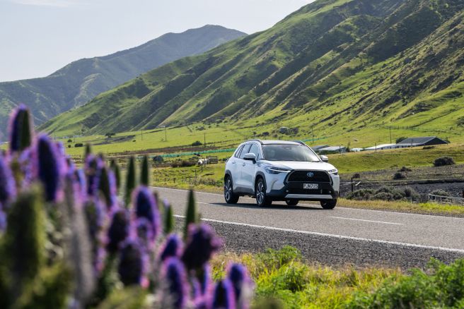 White Toyota hybrid SUV driving along a scenic road with green hills and purple flowers