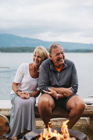 Image of an older couple sitting on a bench with a fire in the foreground and a lake in the background