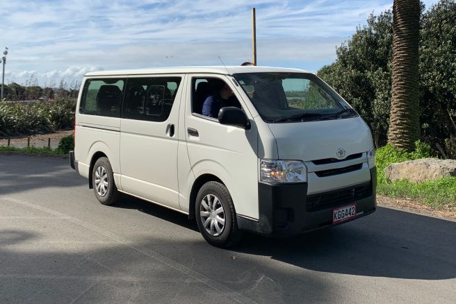 White Toyota Hiace minibus with a driver at the wheel pulling out onto a coastal road lined with flax and palms