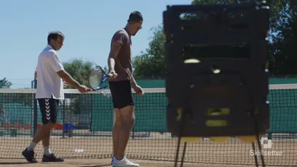 A tennis coach adjusting a player's grip at the net beside a ball machine
