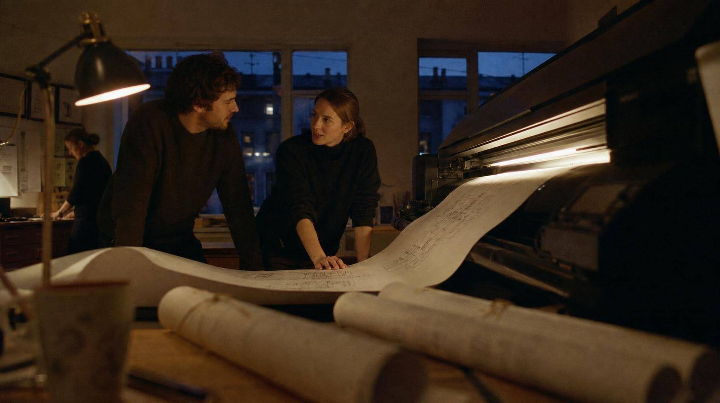Two architects either side of a plotter at dusk, a wide drawing feeding out