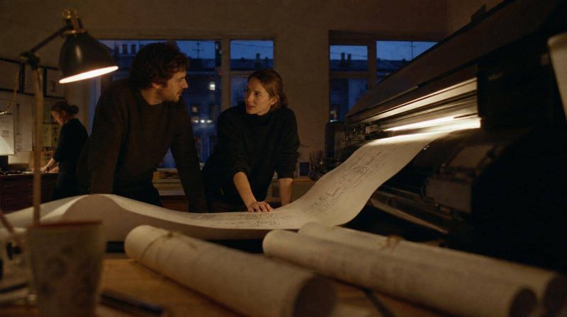 Two architects either side of a plotter at dusk, a wide drawing feeding out