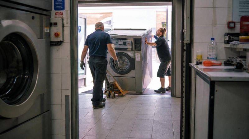 Two people guiding a new industrial washer on a pallet truck through a doorway