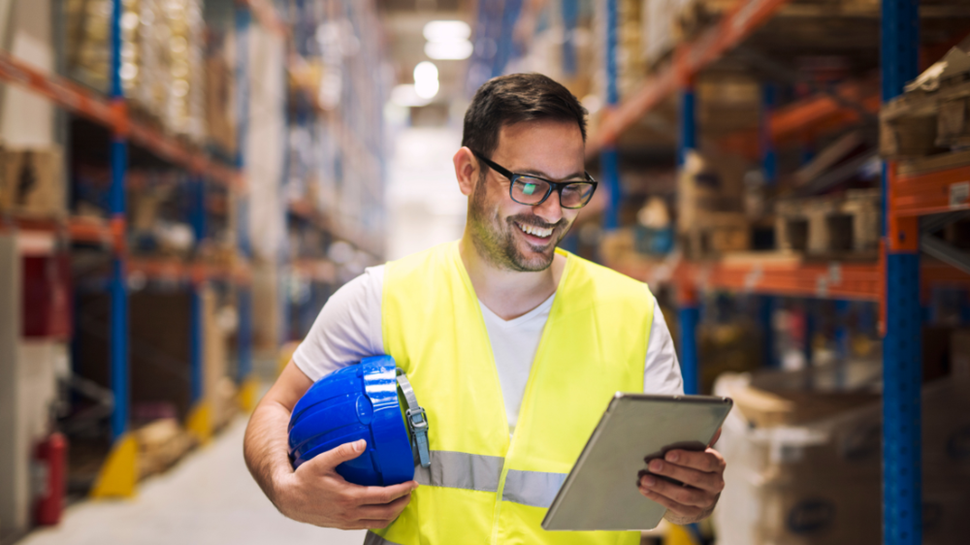Homme en gilet de sécurité jaune tenant un casque bleu et une tablette dans un entrepôt, illustrant l’utilisation du TMS SCM pour optimiser la gestion logistique.