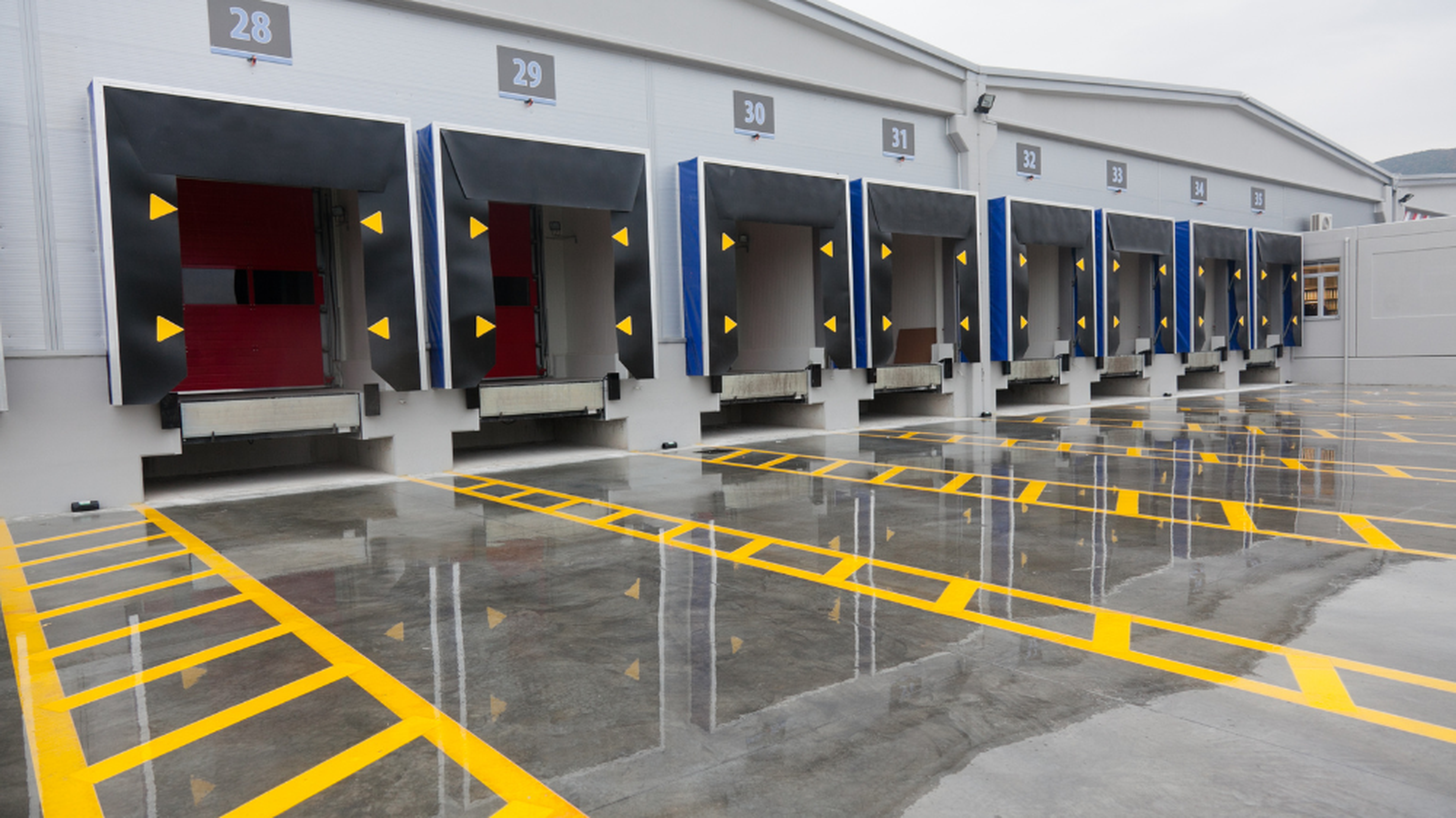 Quais de chargement avec portes rouges, auvents noirs et marquages de sécurité jaunes sur sol béton humide, illustrant la logistics management et l’exécution des flux logistiques.