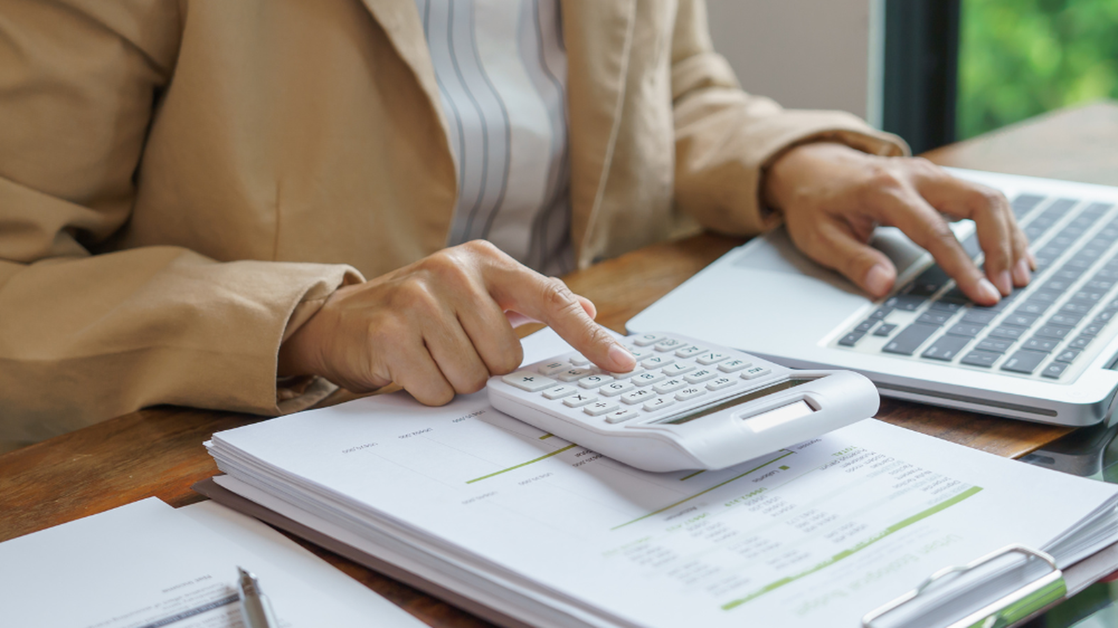 Persona usando una calculadora y un portátil sobre un escritorio con documentos financieros, vestida con una chaqueta beige, analizando datos con un sistema de gestión del transporte (TMS).