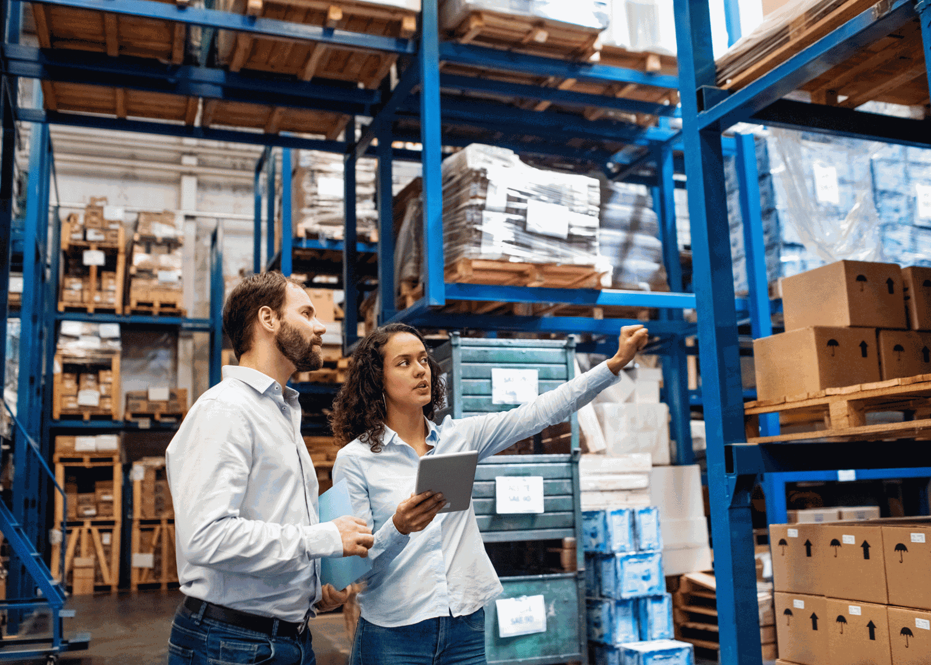 Deux personnes dans un entrepôt, dont l’une tient une tablette, examinent des cartons empilés sur des étagères métalliques bleues, dans un contexte de gestion optimisée grâce à une solution TMS.