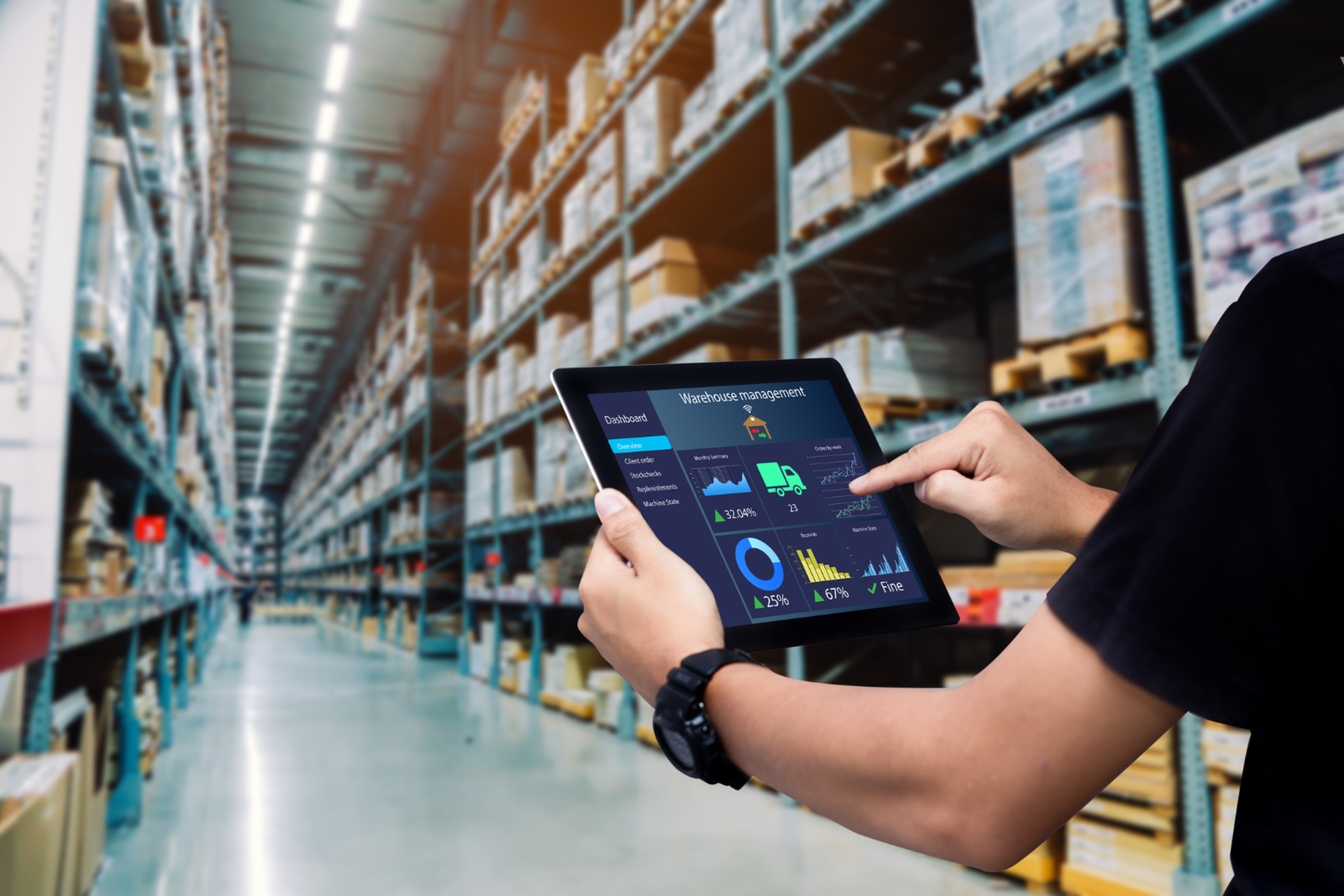 Persona utilizando una tablet para gestionar el inventario en un gran almacén, rodeada de estanterías con cajas y productos, gracias a un sistema de gestión del transporte (TMS).