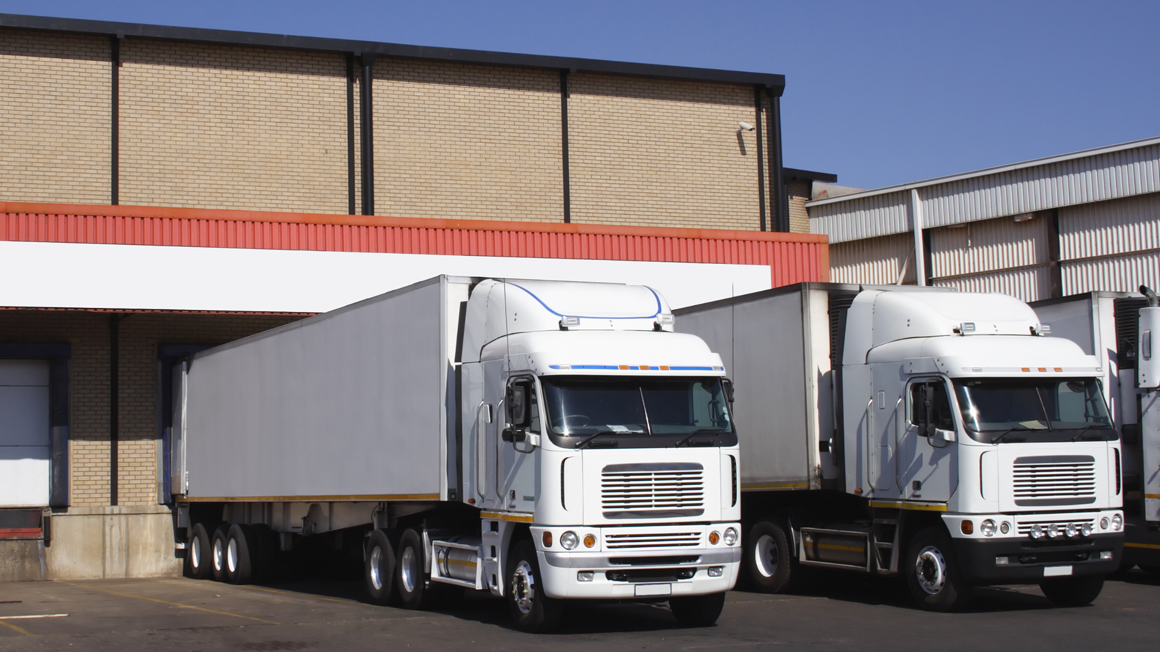 Deux camions blancs stationnés à un quai de chargement devant un entrepôt en briques, illustrant la logistique avec WMS TMS.