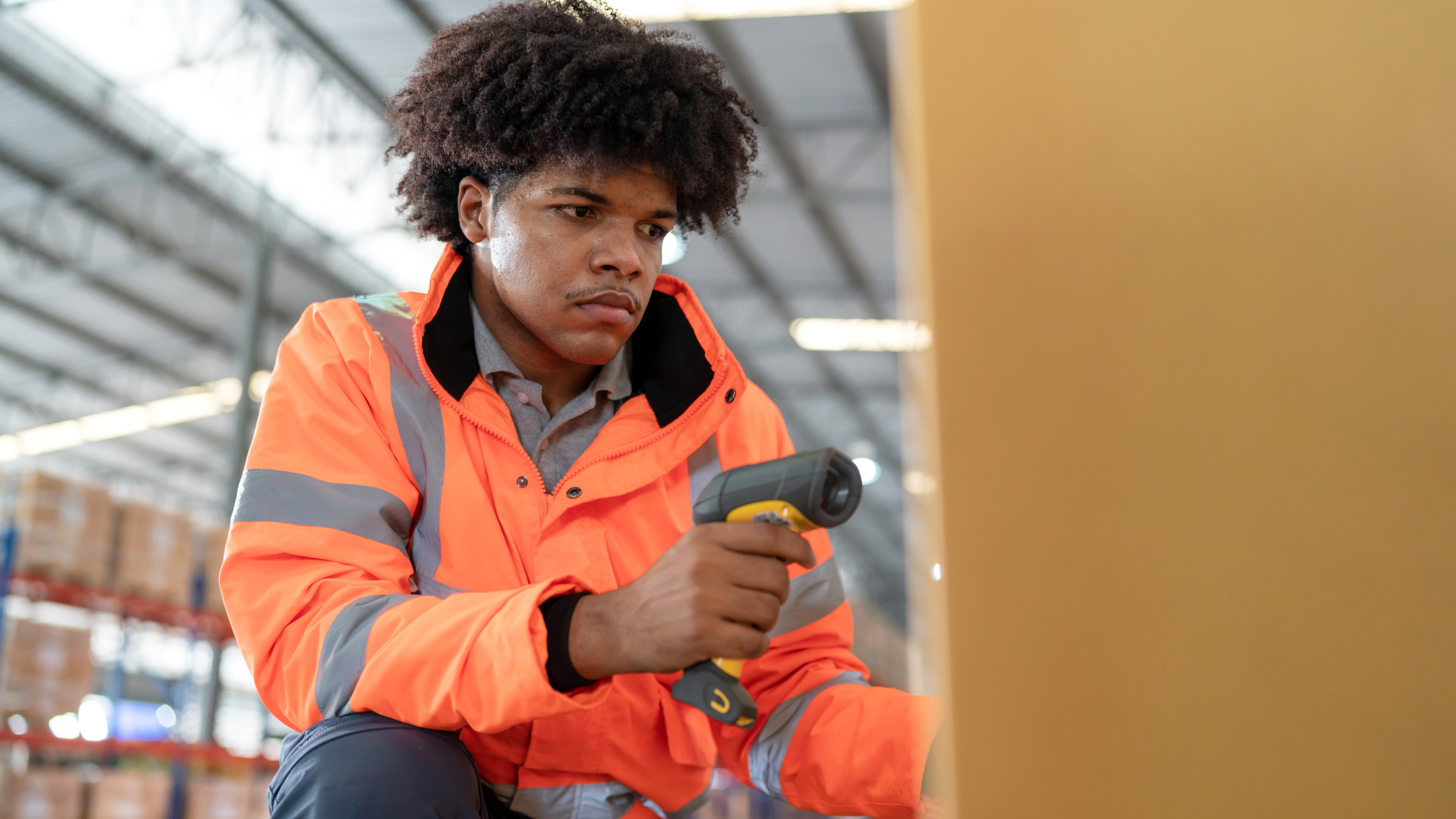 Un opérateur en gilet de sécurité orange scanne un colis à l’aide d’un lecteur de codes-barres dans un entrepôt, illustrant la traçabilité logistique des marchandises.