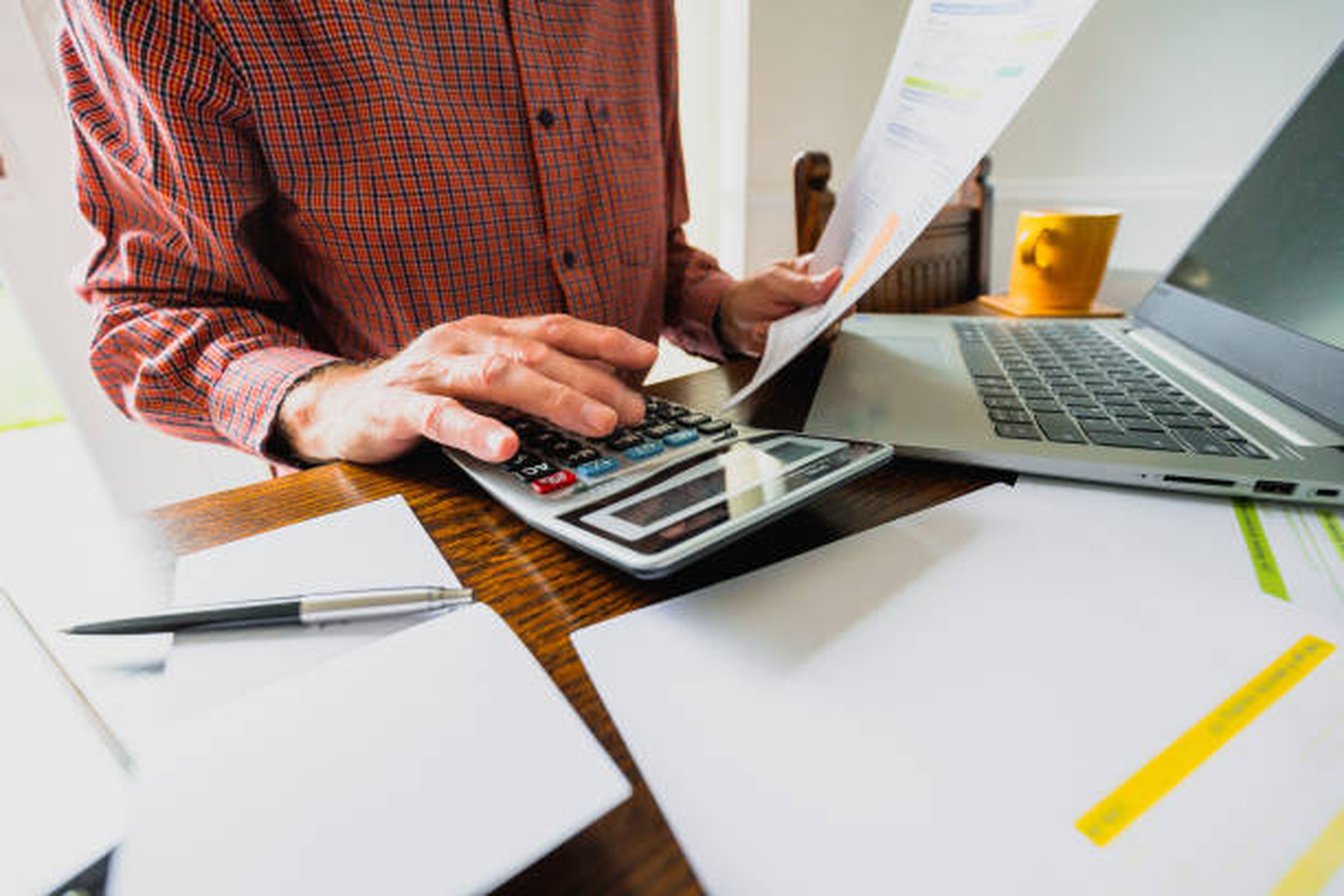 Personne utilisant une calculatrice et analysant un document de coûts liés au TMS transport sur un bureau avec ordinateur portable, stylos, dossiers et tasse en arrière-plan.