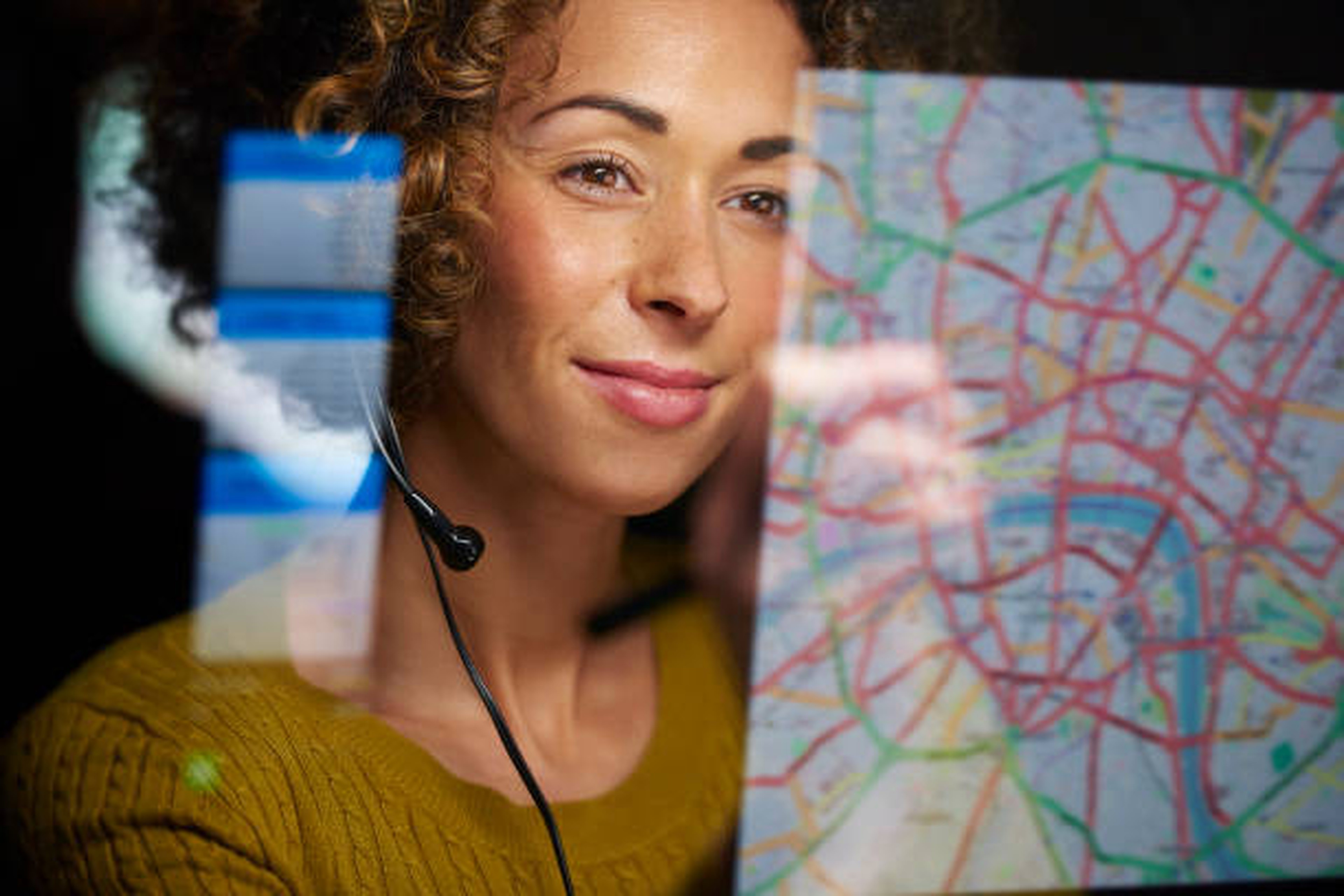 Mujer con auriculares mirando un mapa digital en una pantalla, con un suéter amarillo y expresión pensativa, supervisando un plan de transporte.