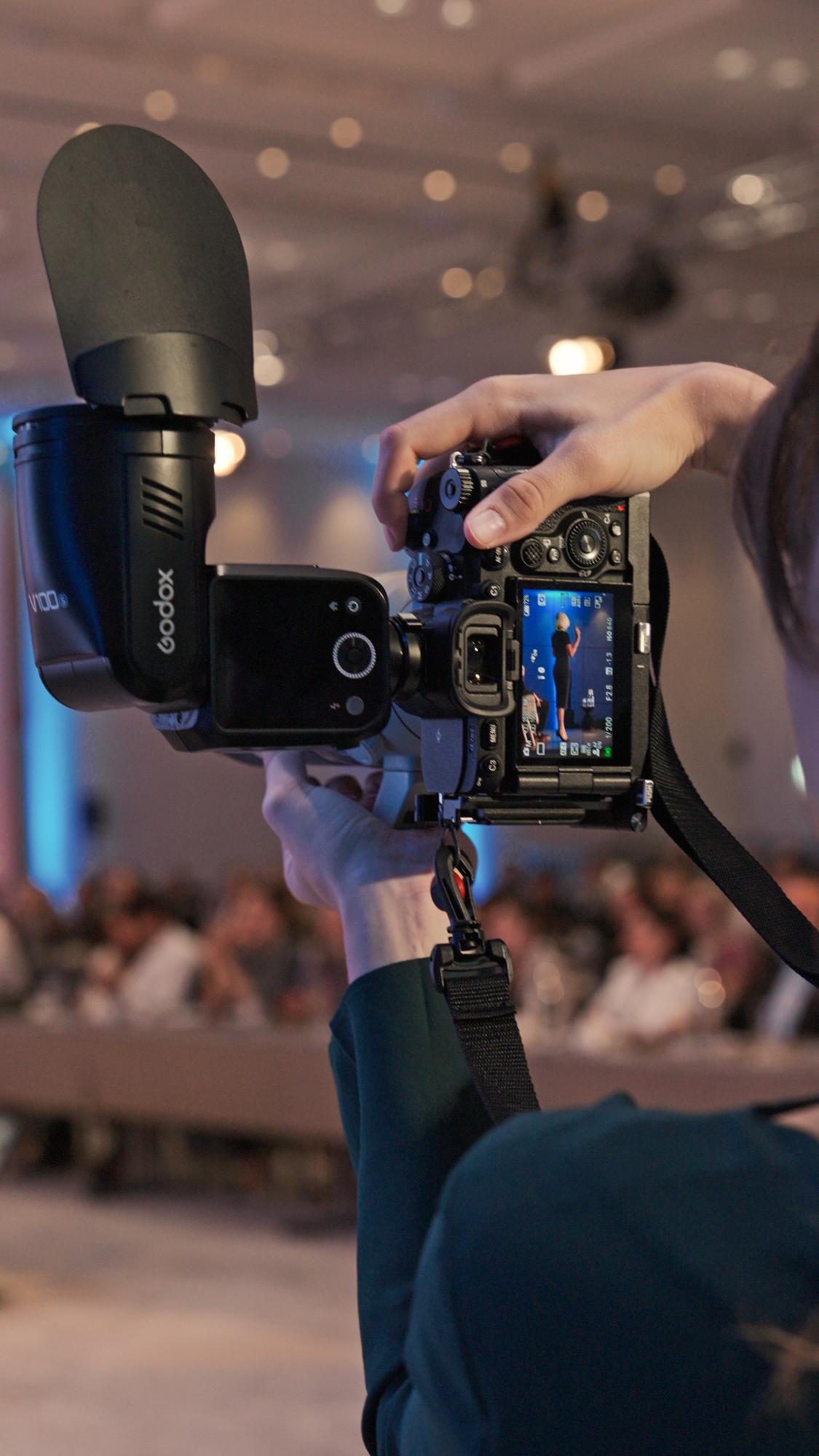 Fotografin mit Kamera und Aufsteckblitz fotografiert eine Rednerin bei einem Kongress
