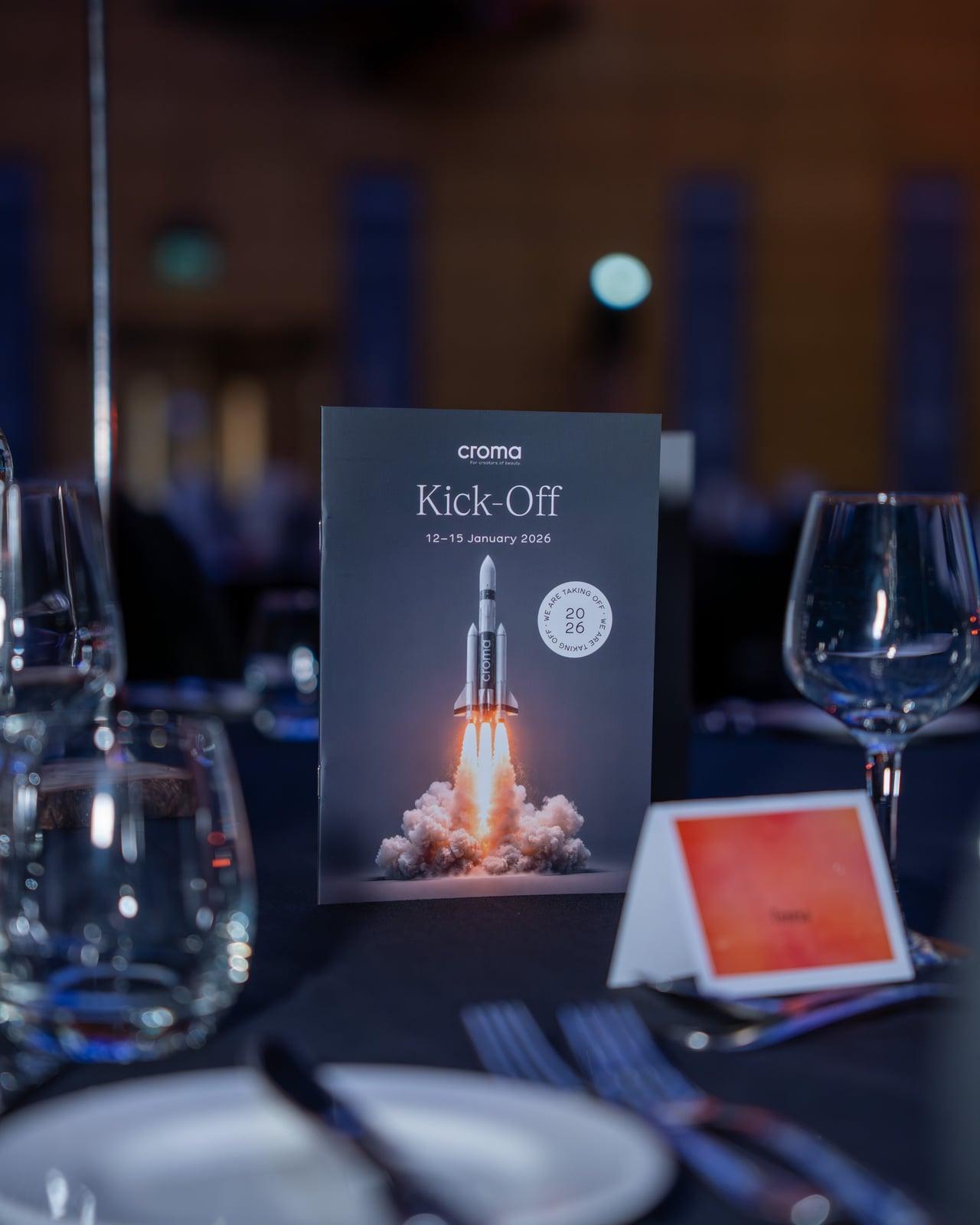 Tischaufsteller „croma Kick-off“ mit Raketenmotiv zwischen Weingläsern