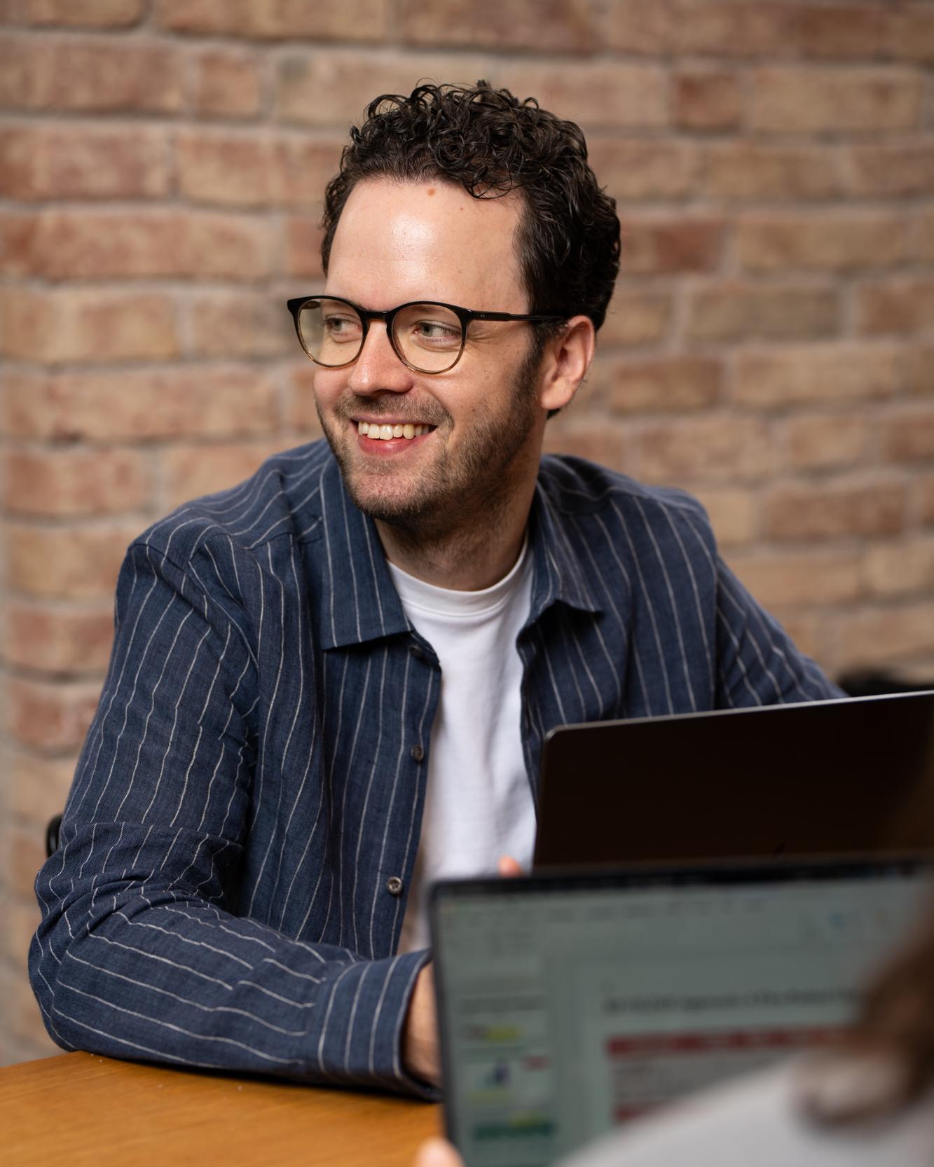 Lächelnder Mann mit Brille sitzt mit Laptops an einem Holztisch vor einer Ziegelwand