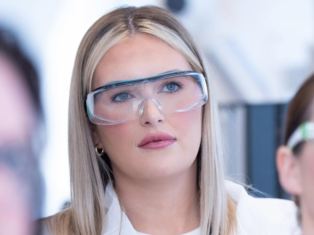 Teilnehmerin mit Schutzbrille und weißem Kittel hört bei einer Laborführung zu