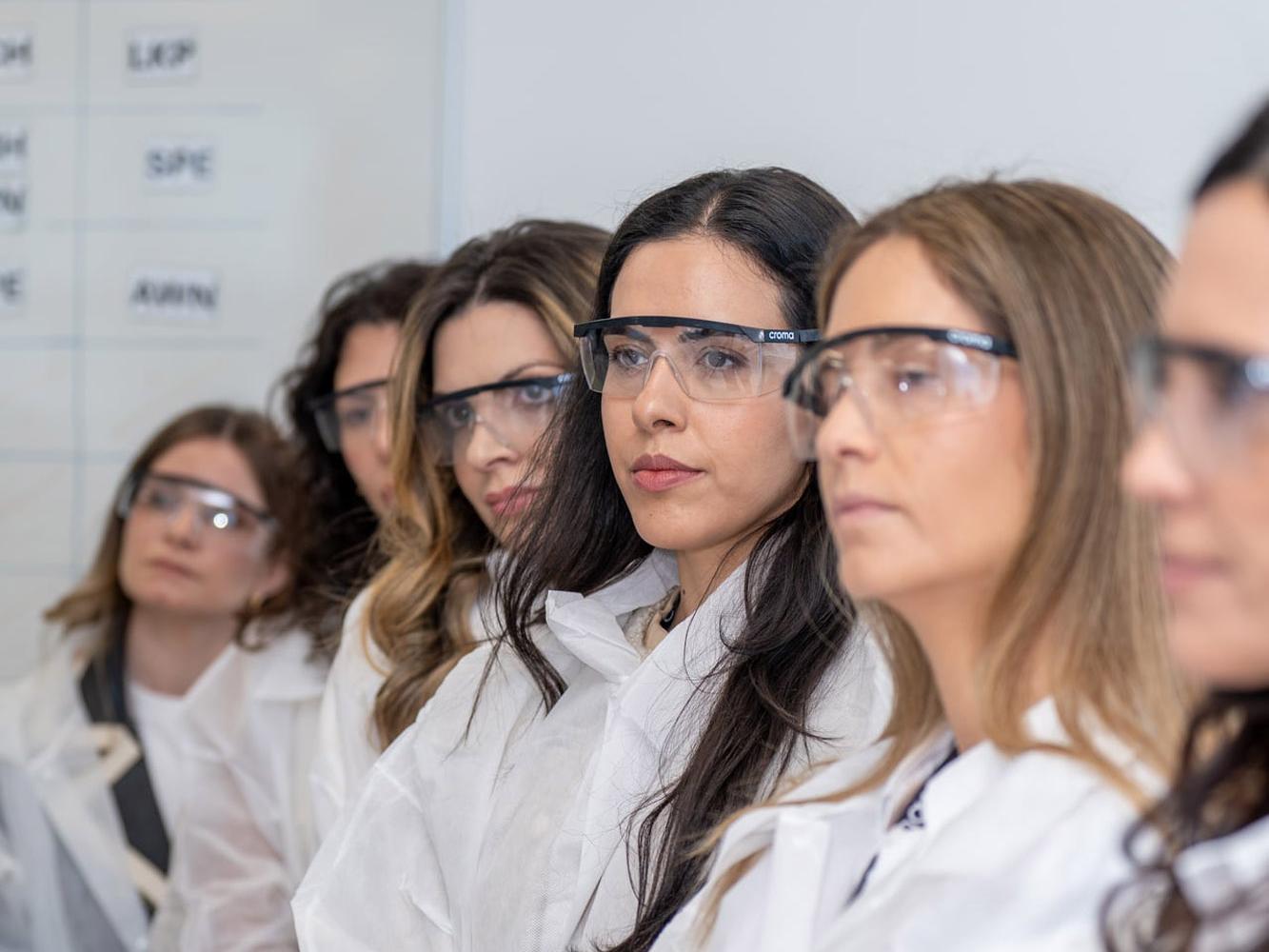 Frauen in weißen Laborkitteln und Schutzbrillen stehen in einer Reihe bei einer Werksführung