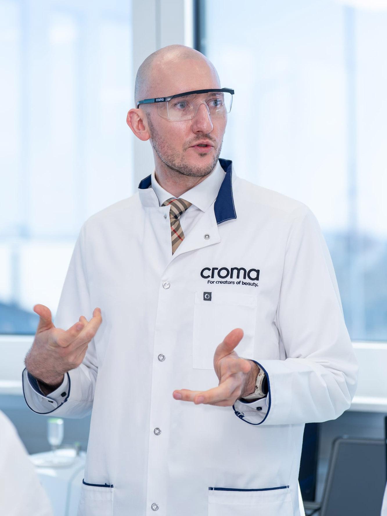Mann in Croma-Laborkittel und Schutzbrille erklärt etwas mit beiden Händen