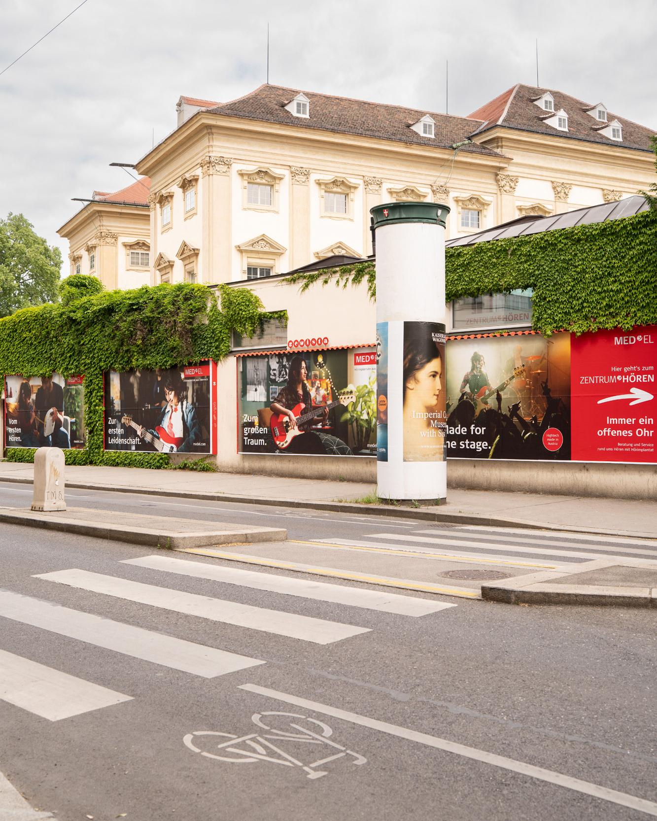 Litfaßsäule und Fassadenwerbung der MED-EL-Kampagne an einer Wiener Straßenkreuzung