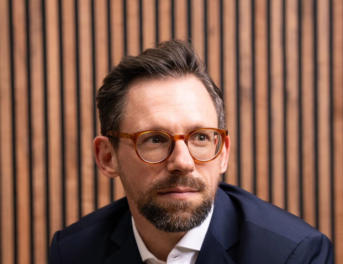 Mann im dunklen Anzug mit Brille vor einer Holzlamellenwand