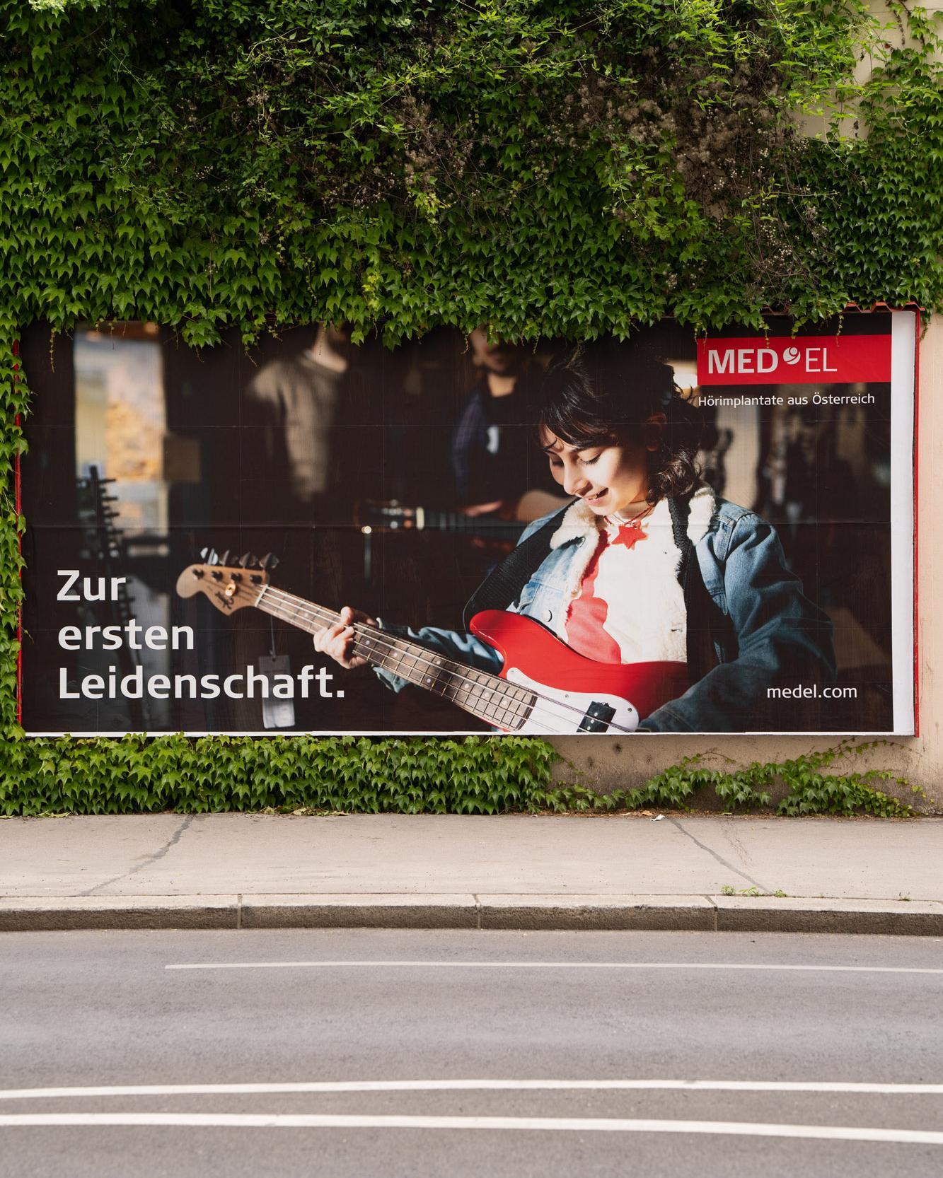 MED-EL-Plakat an einer Hauswand: Mädchen mit E-Gitarre, Schriftzug „Zur ersten Leidenschaft“