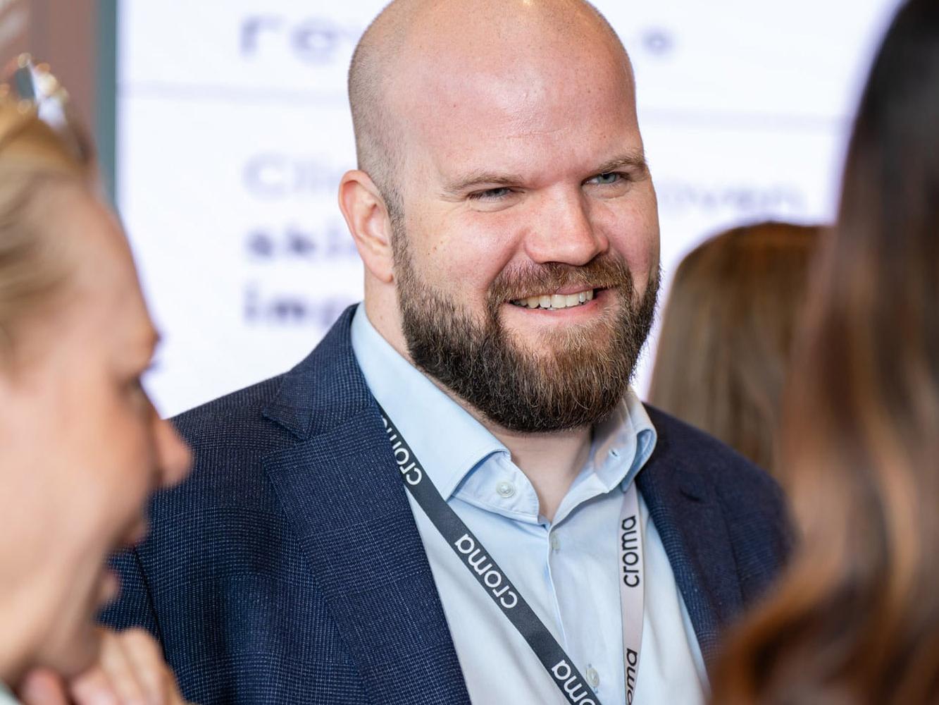 Lächelnder Mann mit Bart und Croma-Umhängeband im Gespräch bei einem Kongress