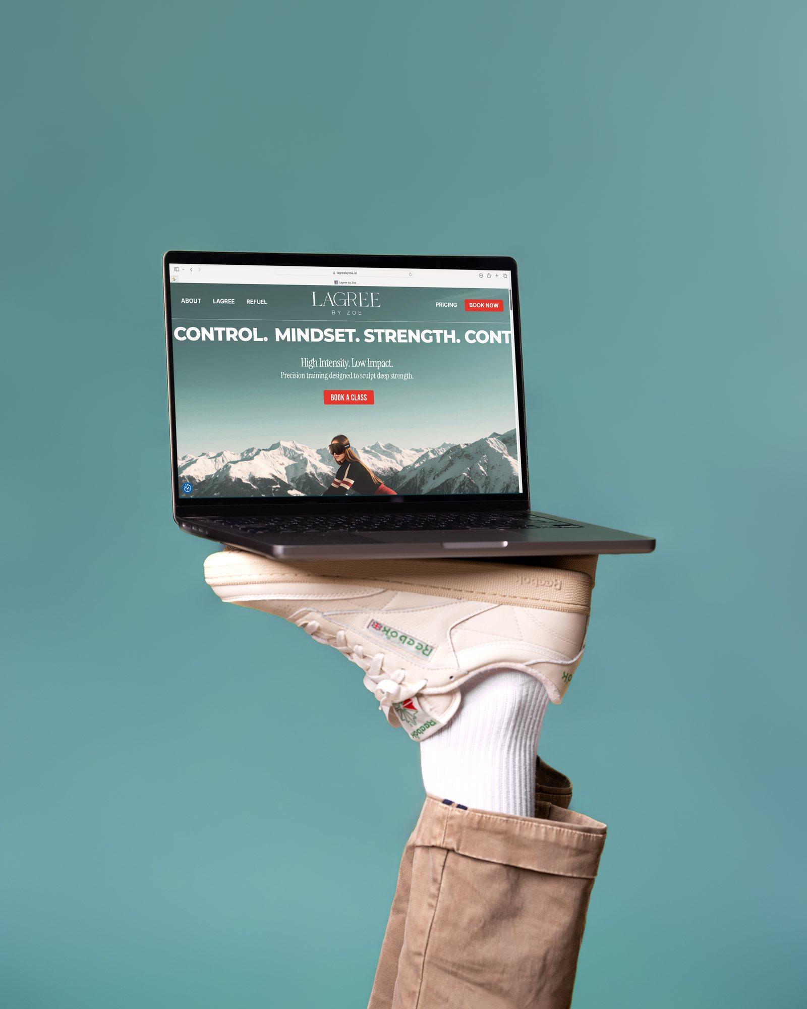 Laptop mit der Website von Lagree by Zoe, balanciert auf einem Sneaker vor petrolfarbenem Hintergrund