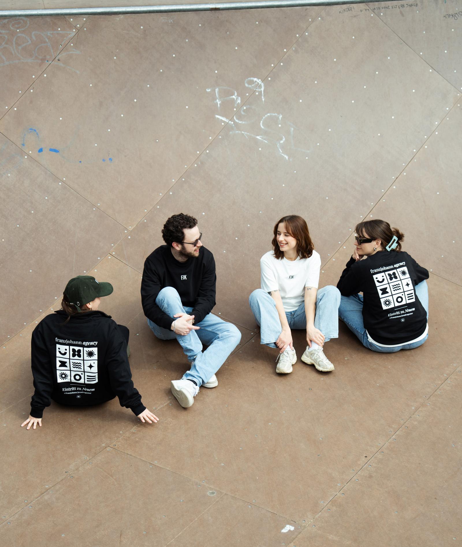 Vier Personen in bedruckten Hoodies sitzen im Kreis auf Asphalt