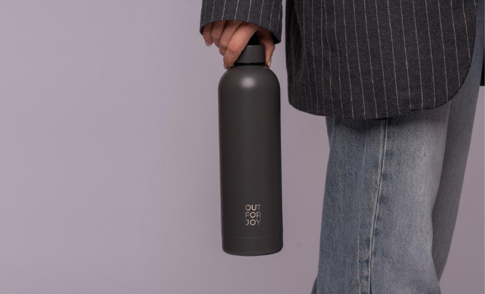 Dunkelgraue Out For Joy Trinkflasche in der Hand, Person in Nadelstreifen-Sakko und Jeans vor fliederfarbener Wand