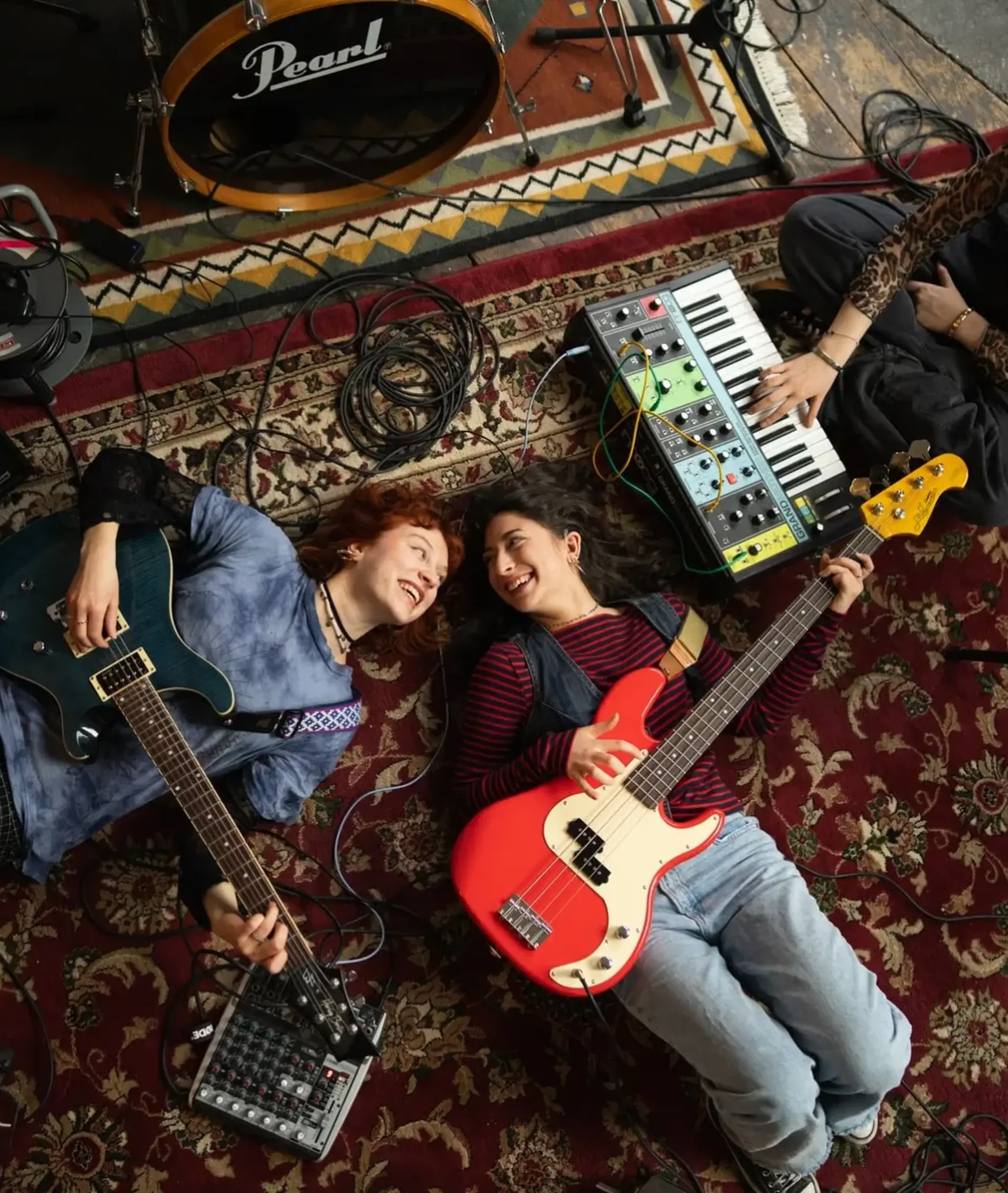 Die Band liegt mit Instrumenten auf einem Teppich im Proberaum, von oben
