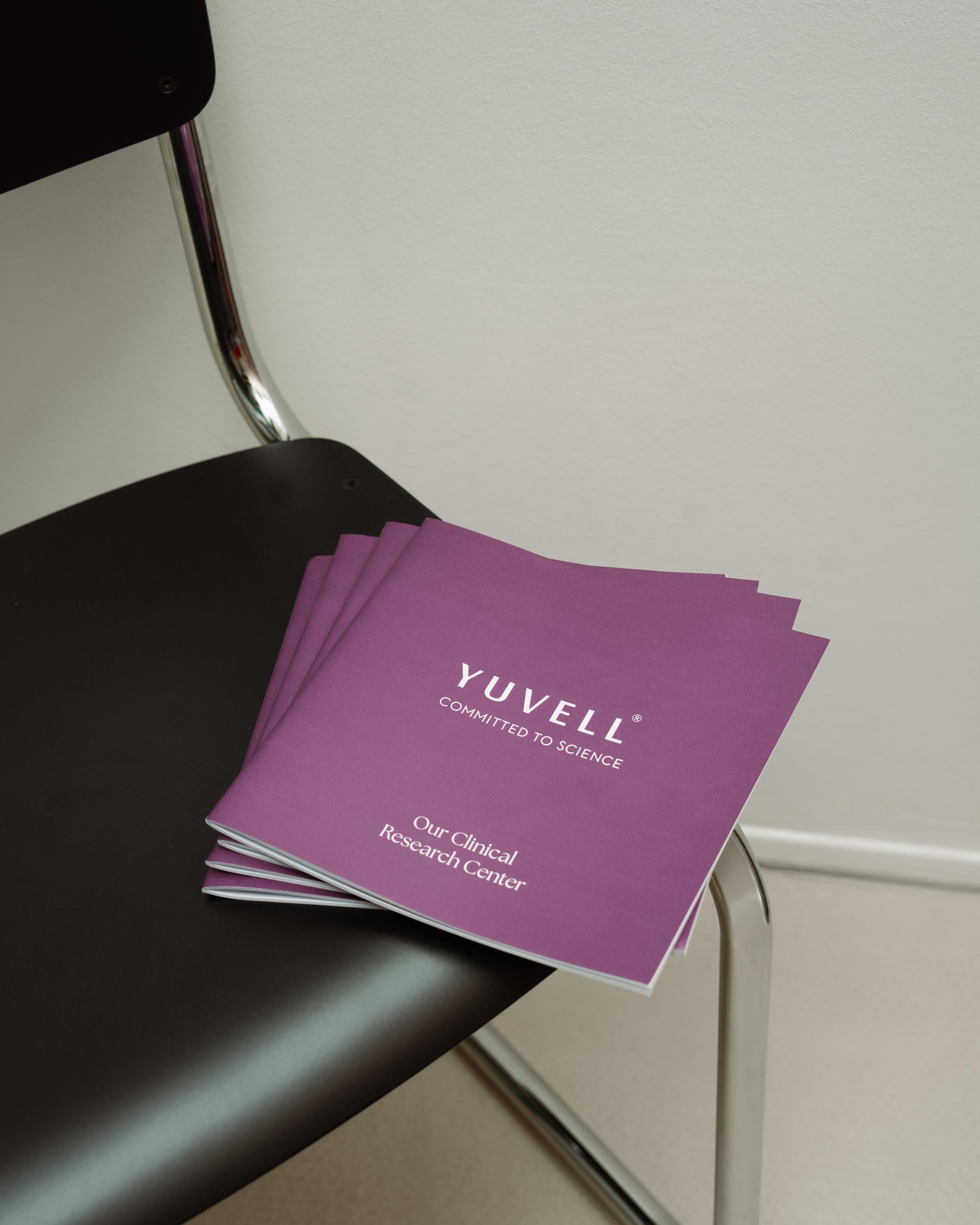 Stapel violetter Broschüren „YUVELL – Our Clinical Research Center“ auf einem Stuhl