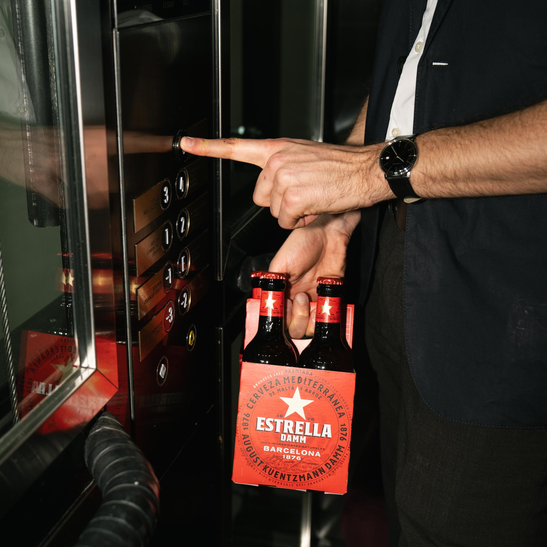 Hand drückt einen Aufzugknopf, daneben Estrella-Bierflaschen