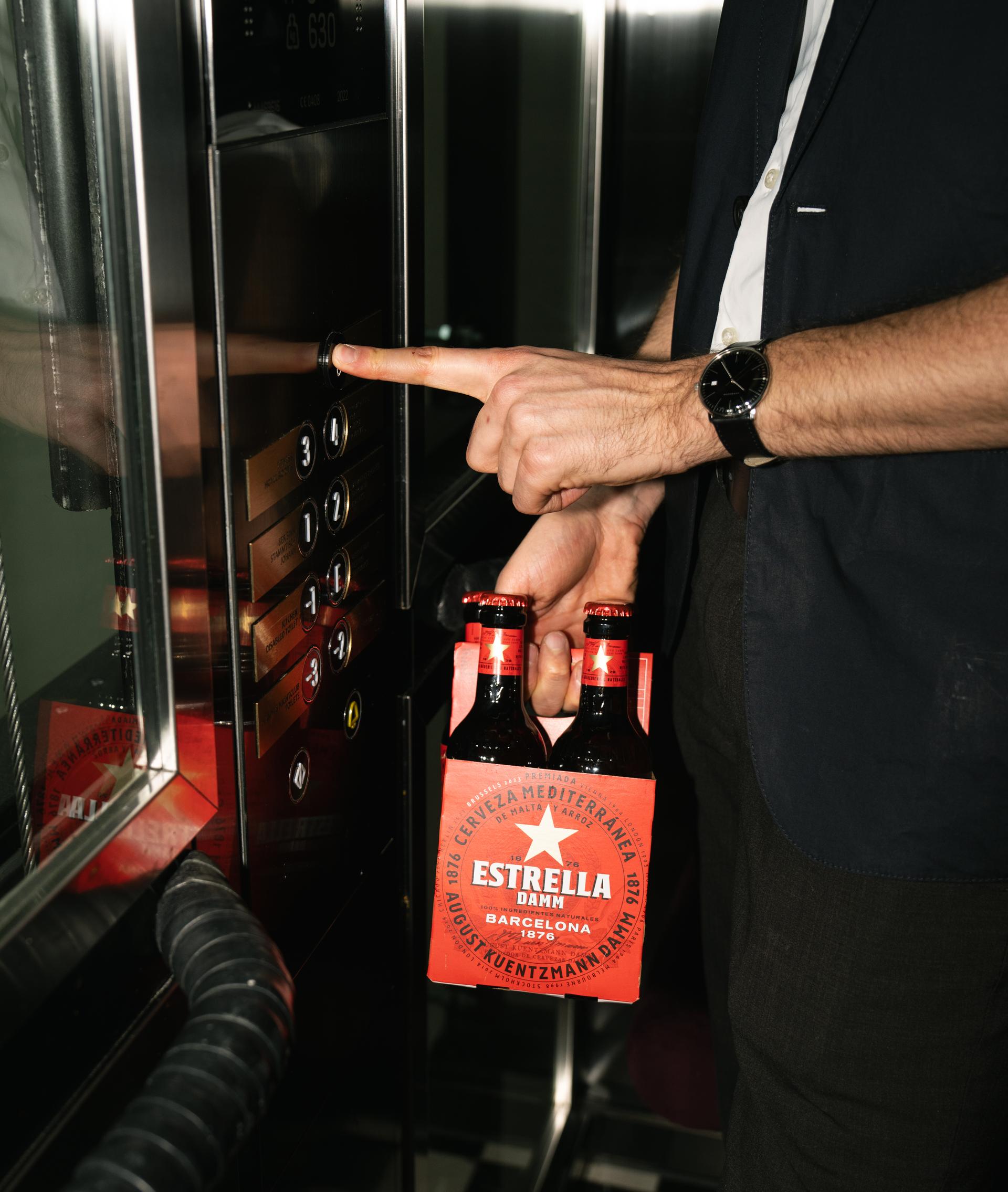 Hand drückt einen Aufzugknopf, daneben zwei Estrella-Bierflaschen
