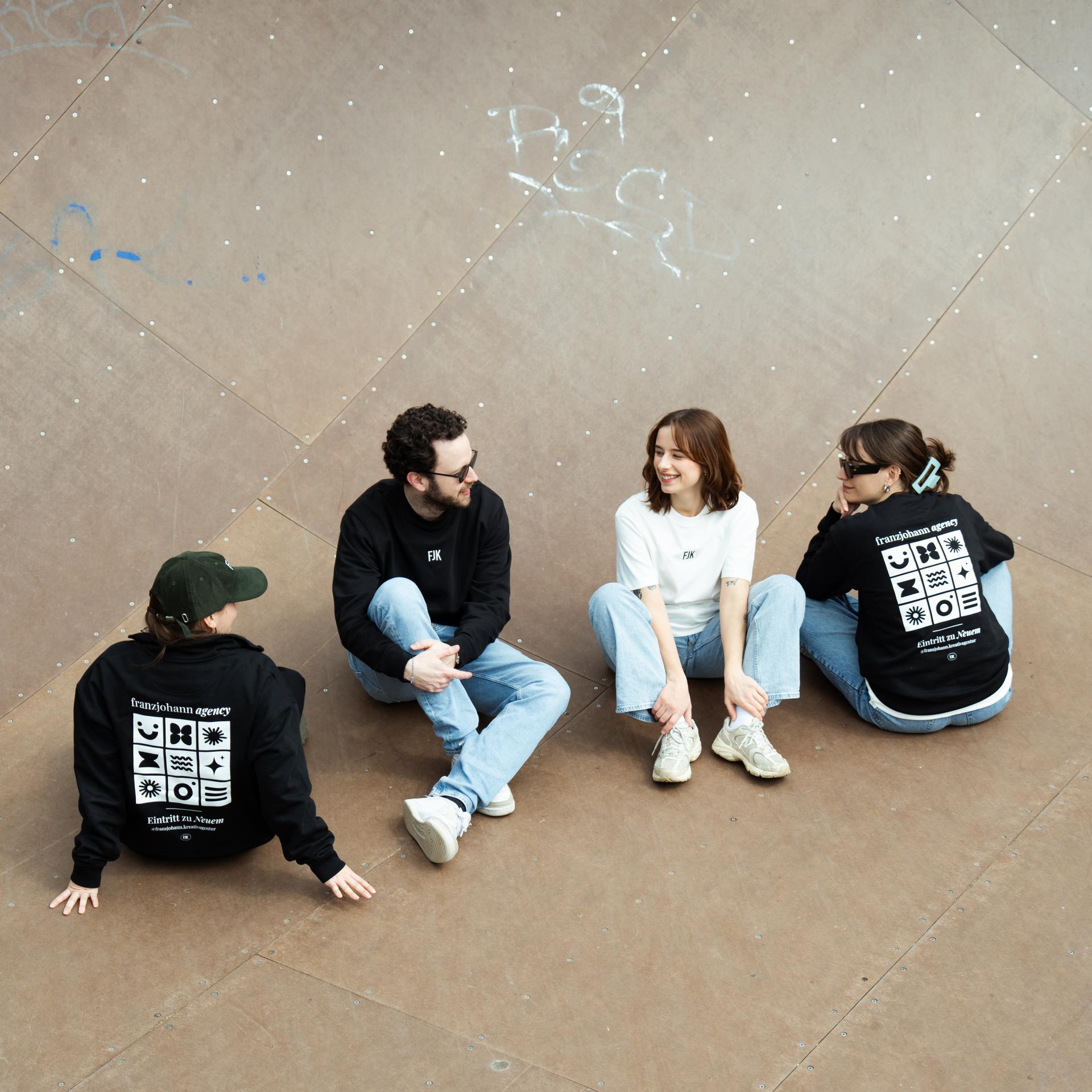 Vier Personen in bedruckten Hoodies und Shirts sitzen auf Asphalt