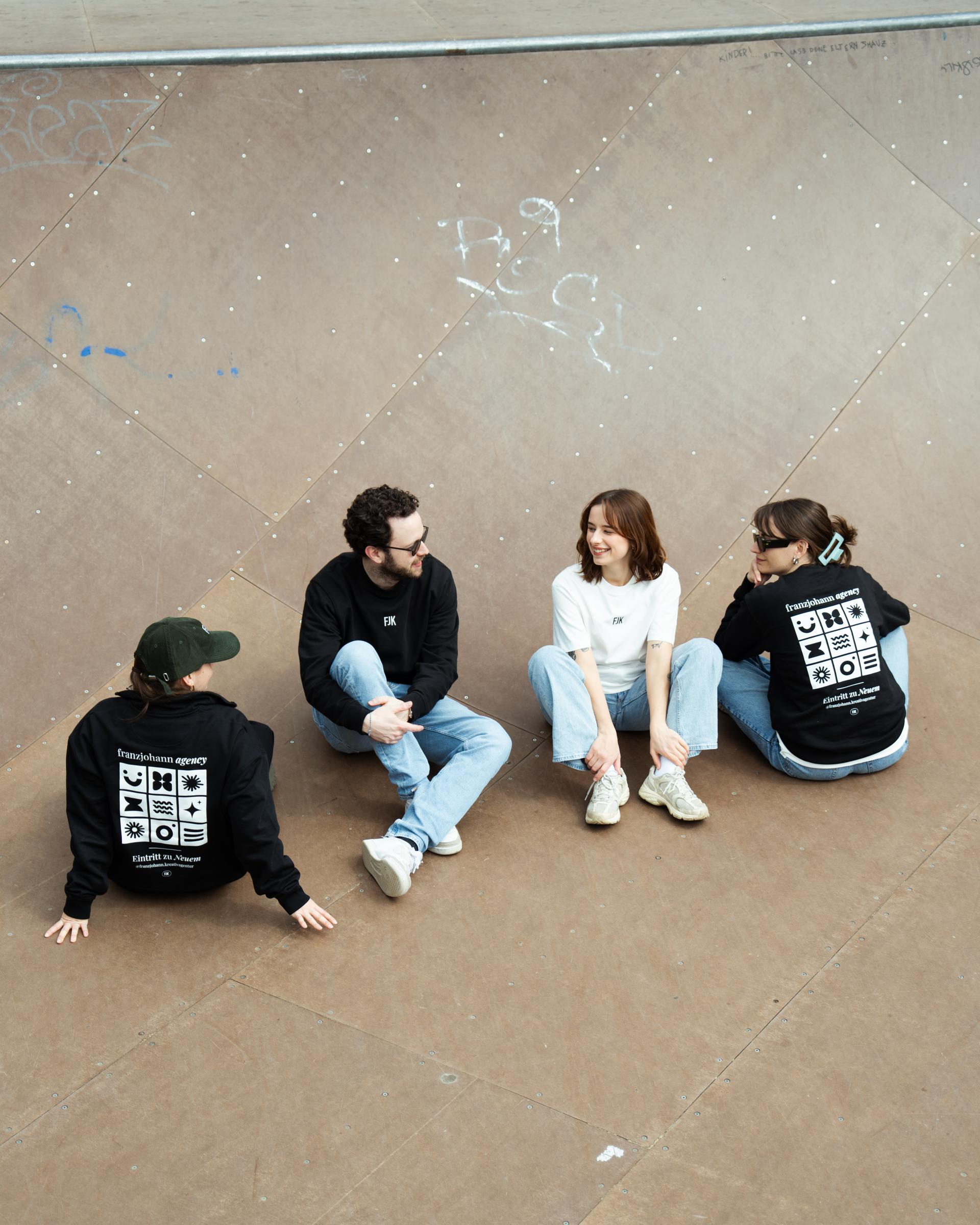 Vier Personen in bedruckten Hoodies und Shirts sitzen auf Asphalt