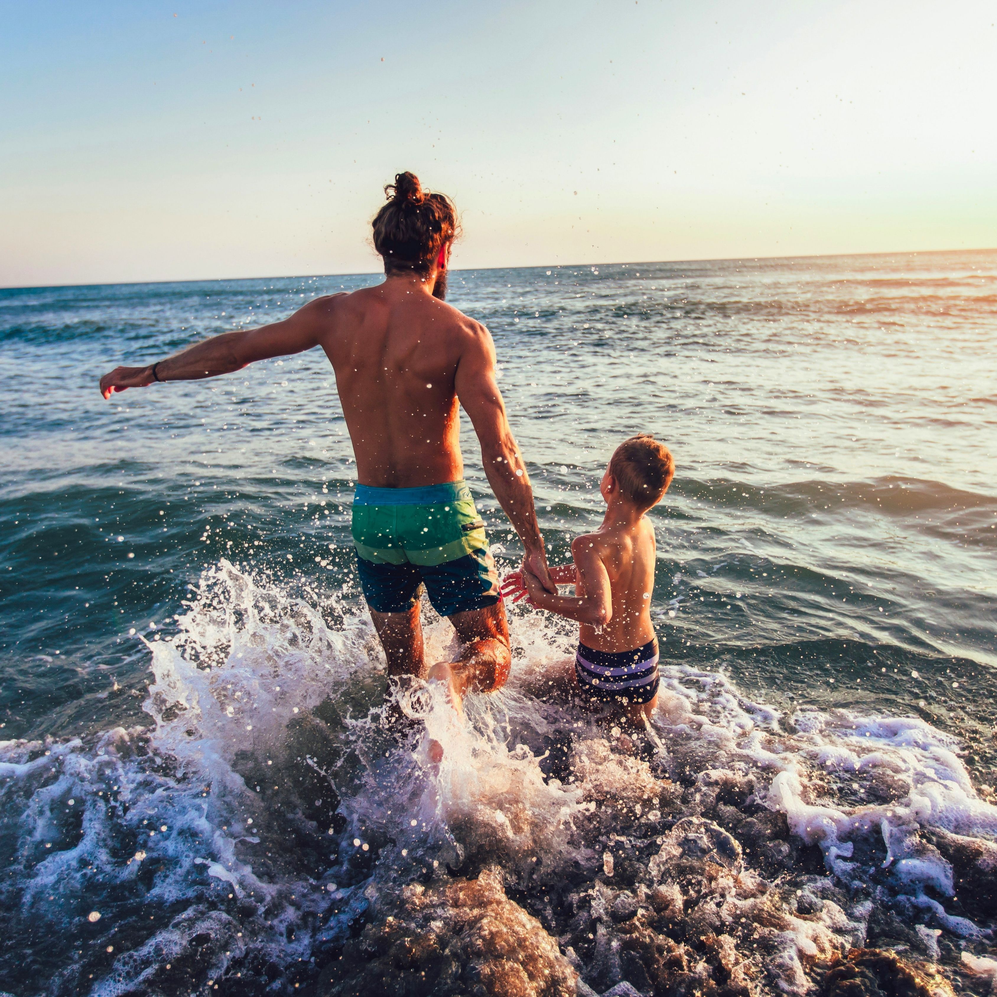 A father and young child hold hands as they run into the ocean, splashing through the waves.