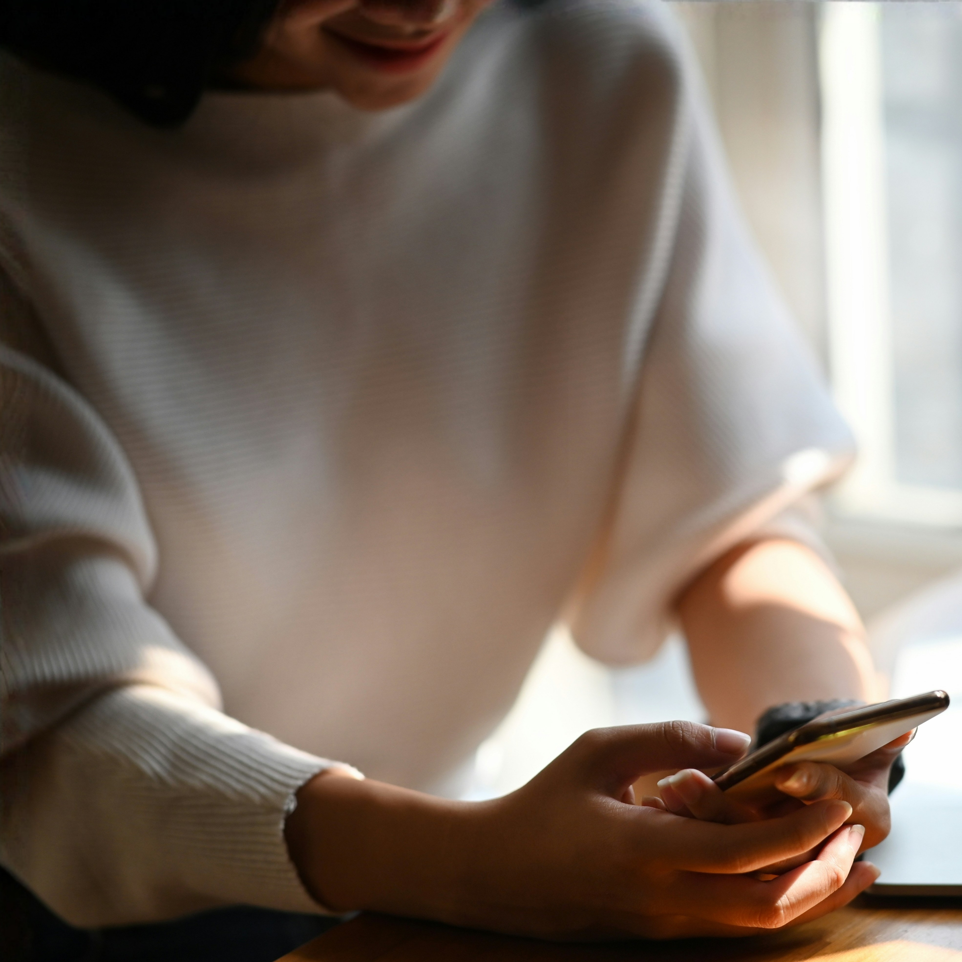 A person in a light sweater uses a smartphone while sitting indoors near a window.