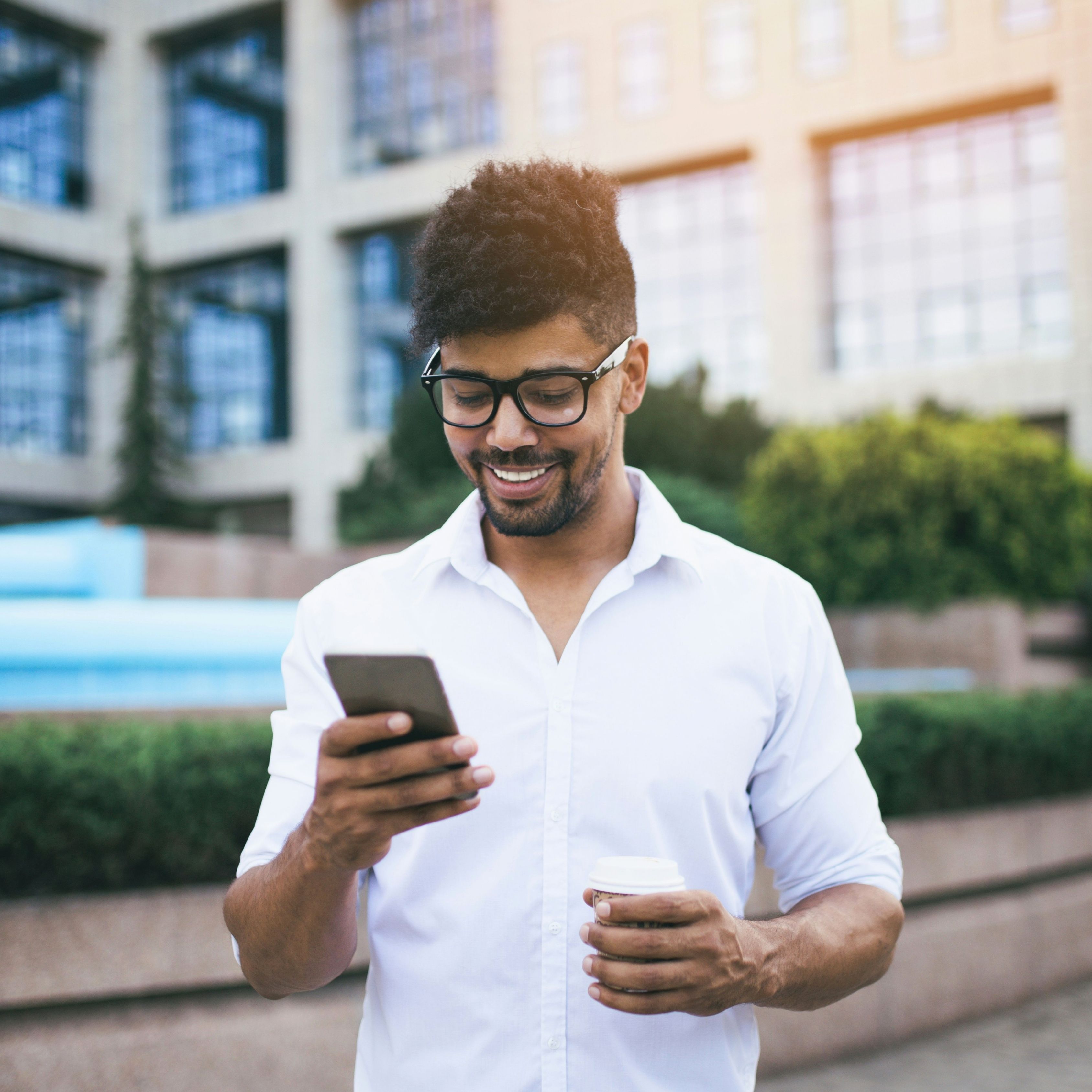 A smiling man looks at his smartphone while holding a coffee cup outdoors.