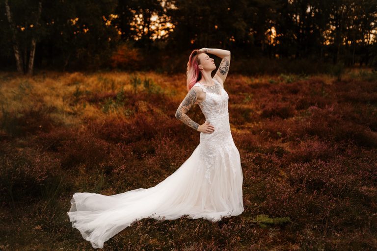 Braut im Spitzenkleid mit langer Schleppe steht in der warm leuchtenden Heide