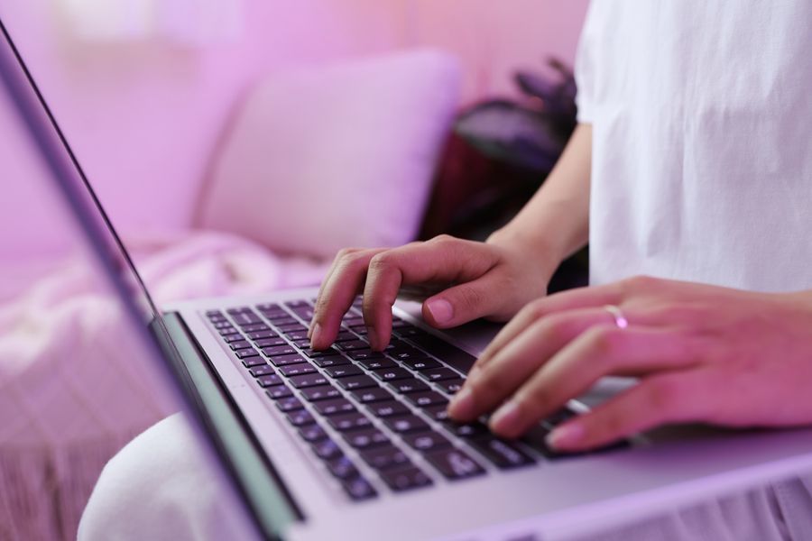 Close-up of a person's hands typing on a laptop keyboard while sitting on a sofa