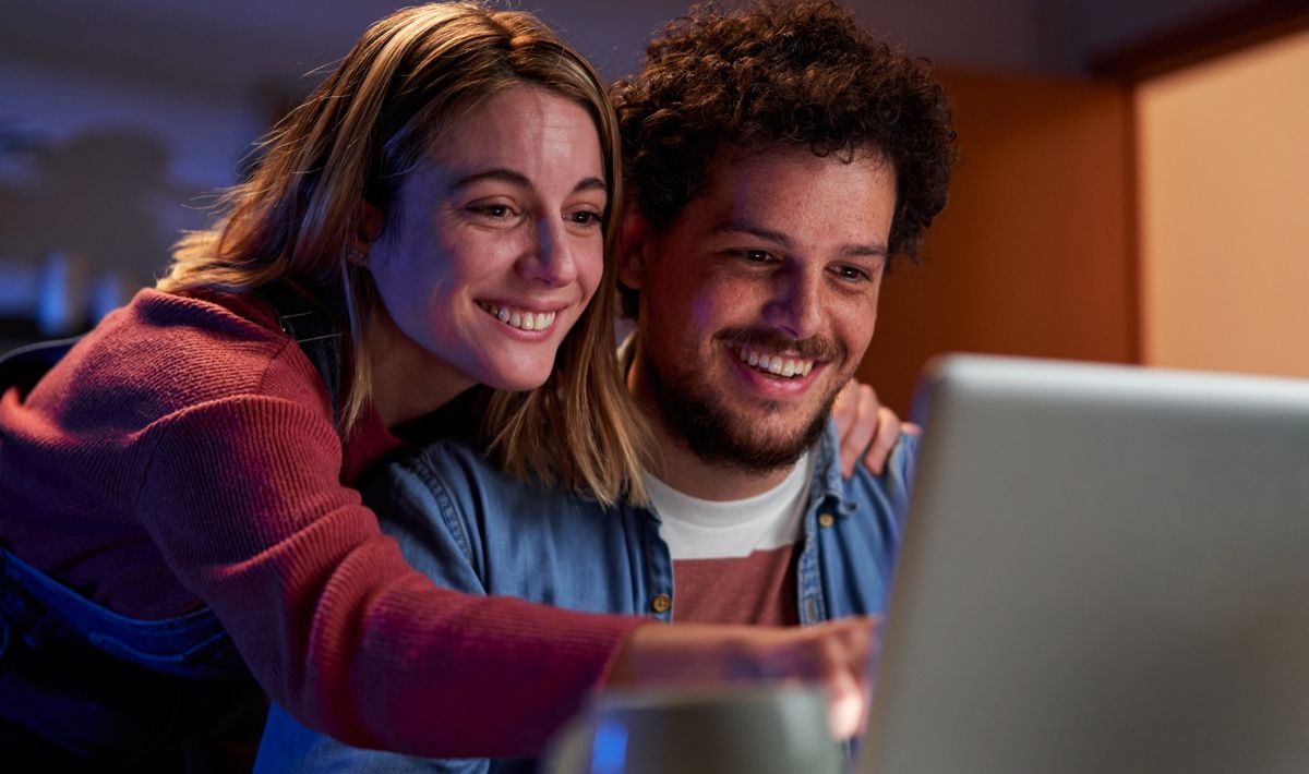 Two people leaning together at a kitchen table, smiling at an open laptop