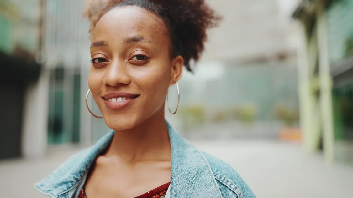 A smiling woman in a denim jacket, standing outdoors in a city