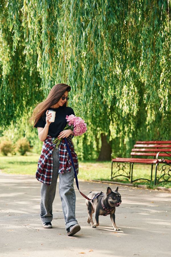 Woman walking a French bulldog along a park path, carrying a coffee cup and a bunch of peonies