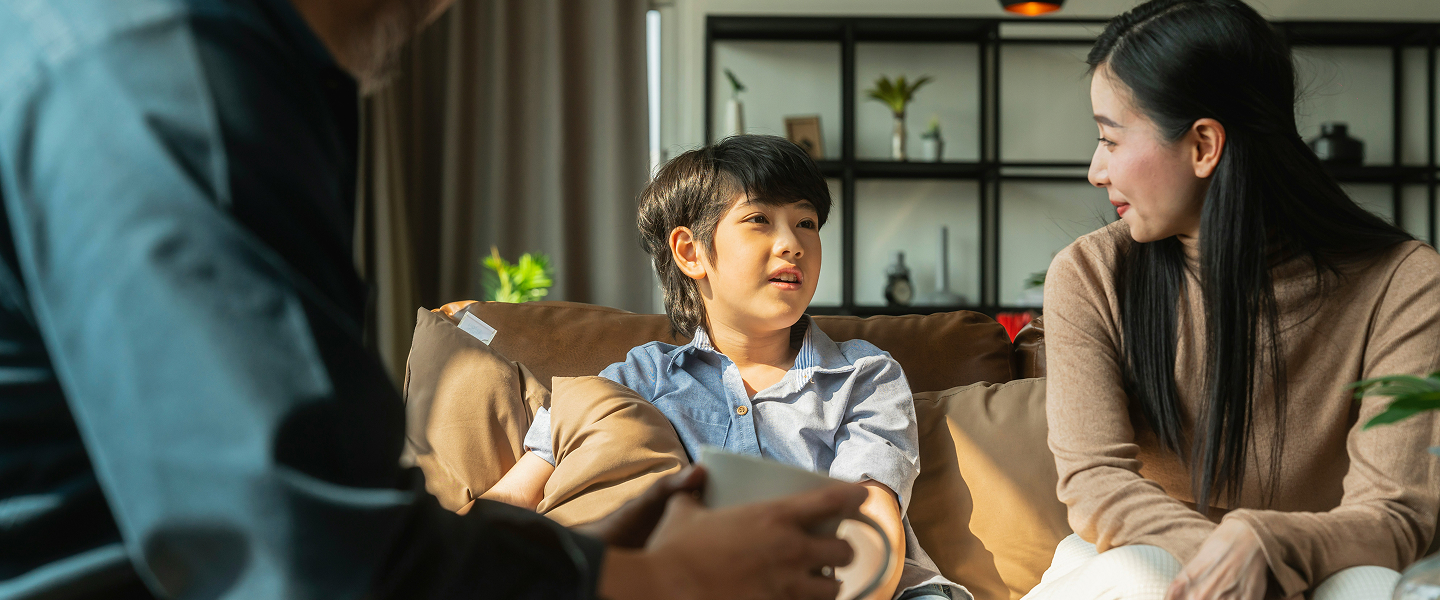A child and a woman sitting together on a couch, talking with an older man who is seated across from them in a living room.