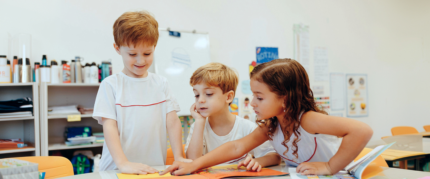 Three young children in white shirts looking at a colourful picture book together at a table in a classroom.
