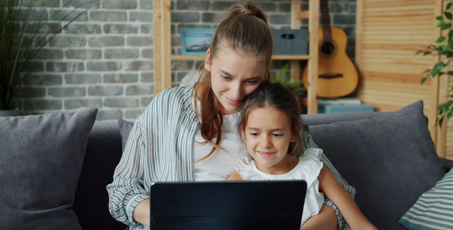A woman and a young girl sitting together on a couch at home, both smiling as they look at an open laptop.