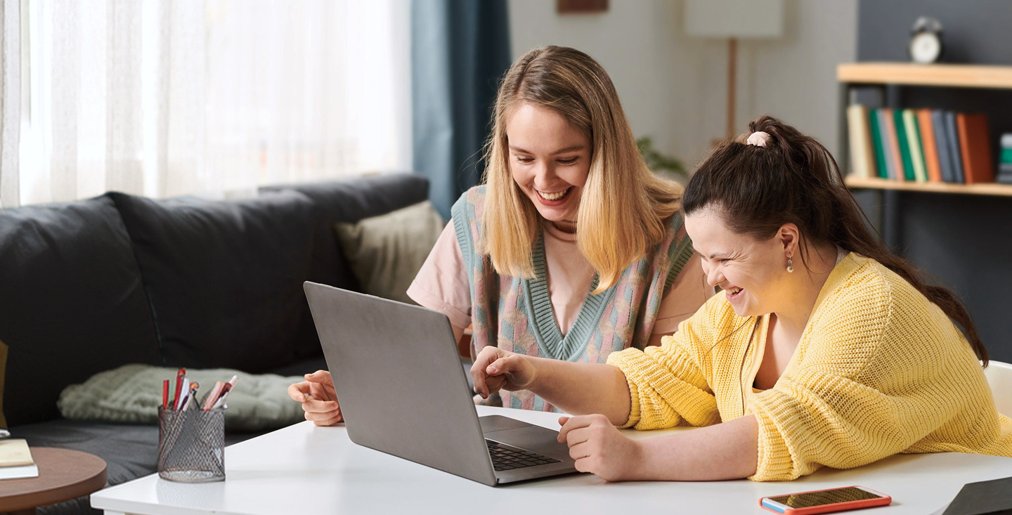 Two young women laughing together at a desk while looking at a laptop in a home setting, with books and stationery nearby.