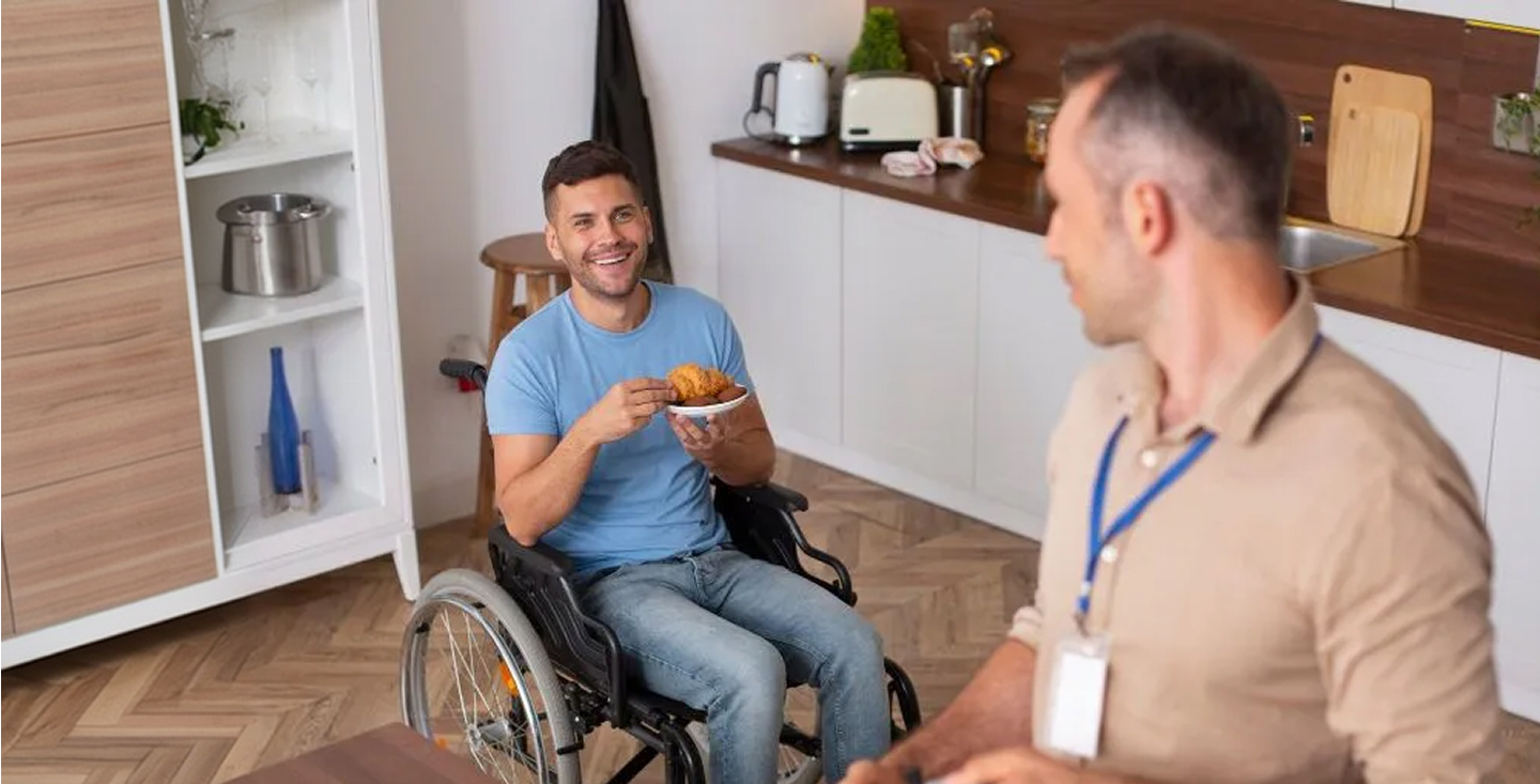A smiling man seated in a wheelchair holding a bowl of food in a kitchen, talking with a man wearing a lanyard who is seated nearby.