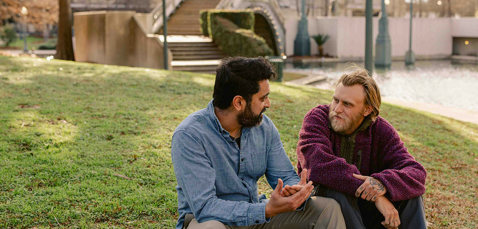 Two men sitting on the grass in a park, having a relaxed conversation.